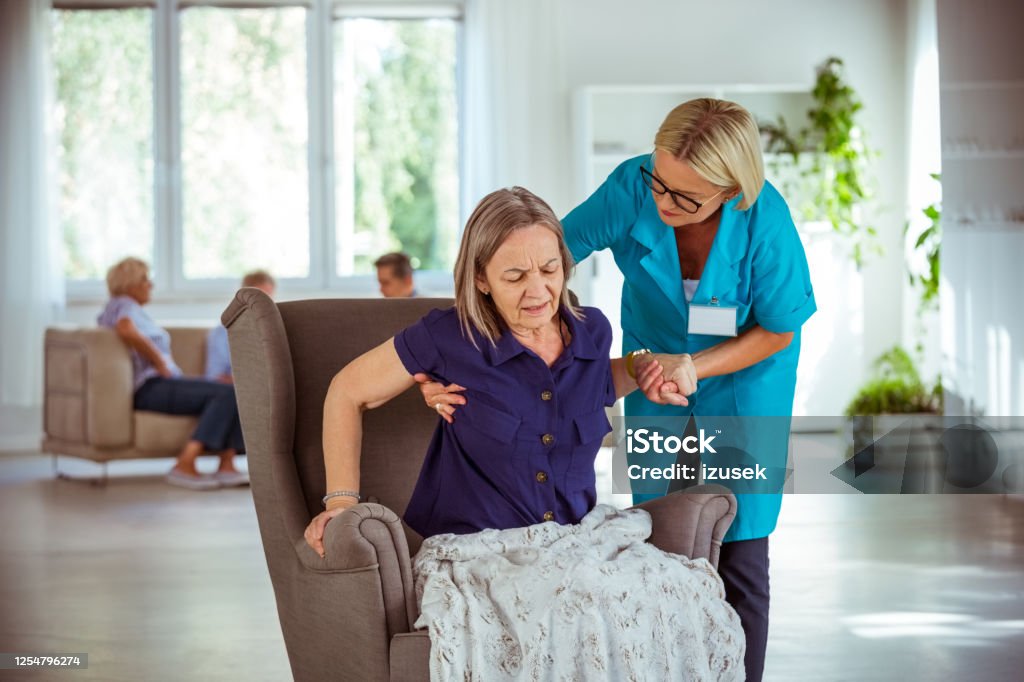 Caregiver helping older lady to stand up Home nurse giving support of old woman to get up from an armchair. They are in retirement home. Parkinson's Disease Stock Photo