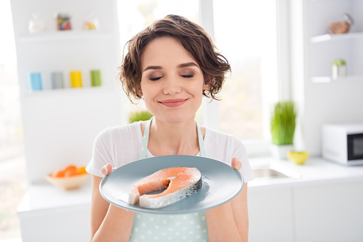 Close up photo of beautiful housewife lady enjoy pleasant smell of fresh raw salmon, fillet steak tasty diet breakfast eyes closed hungry wear apron t-shirt stand modern kitchen indoors