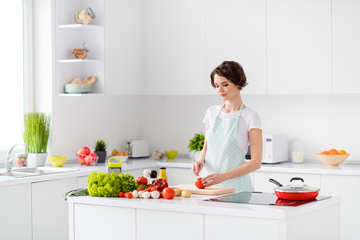 Photo of housewife attractive focused lady arms holding tomato, cutting knife slices enjoy morning cooking tasty yummy dinner wear apron stand modern kitchen indoors