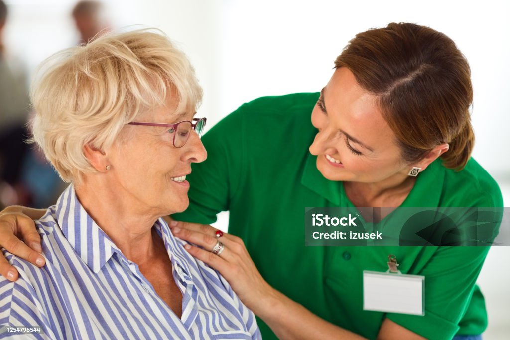 Cheerful home caregiver embracing senior woman Elderly lady in retirement home. Friendly nurse talking with senior woman, sitting in living room together. Embracing Stock Photo