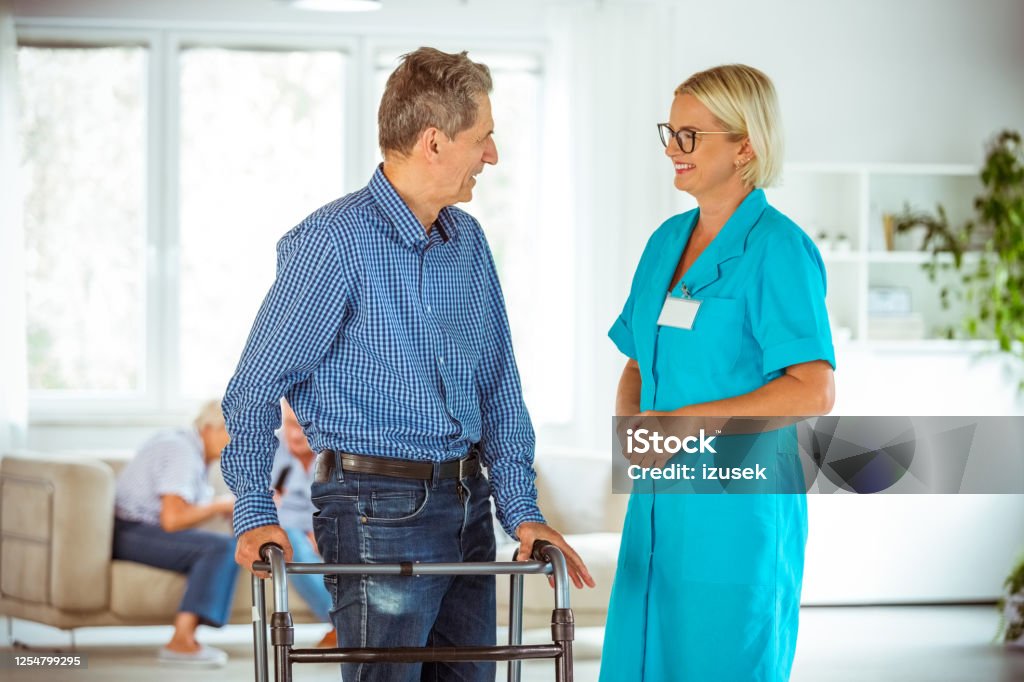 Cheerful nurse talking senior man in retirement house Friendly nurse wearing uniform talking with senior man. Man using walking frame. Nurse Stock Photo