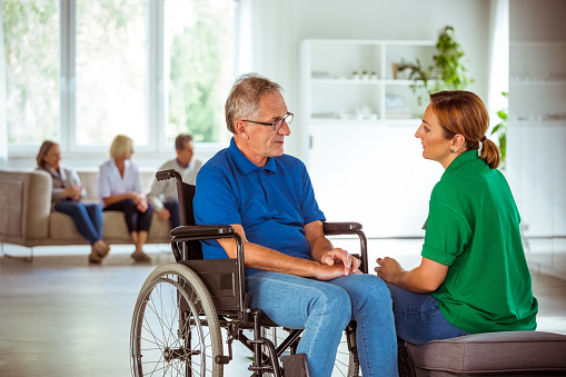 Elderly man in retirement home. Friendly nurse talking with worried senior man sitting in wheelchair.