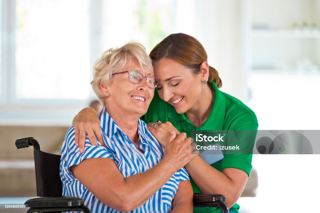 Female social worker supporting senior woman Elderly lady in retirement home. Friendly nurse embracing senior woman sitting in wheelchair. Embracing Stock Photo
