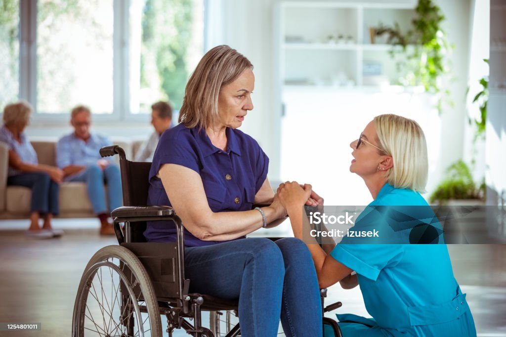 Friendly nurse consoling senior woman Home nurse holding hands of worried senior woman in retirement house. Elderly lady sitting in wheelchair. 70-79 Years Stock Photo