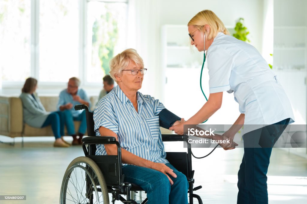 Home nurse checking the blood pressure of the elderly lady Female home nurse measuring the arterial pressure of the senior woman. An elderly lady sitting in wheelchair in a retirement home. 70-79 Years Stock Photo