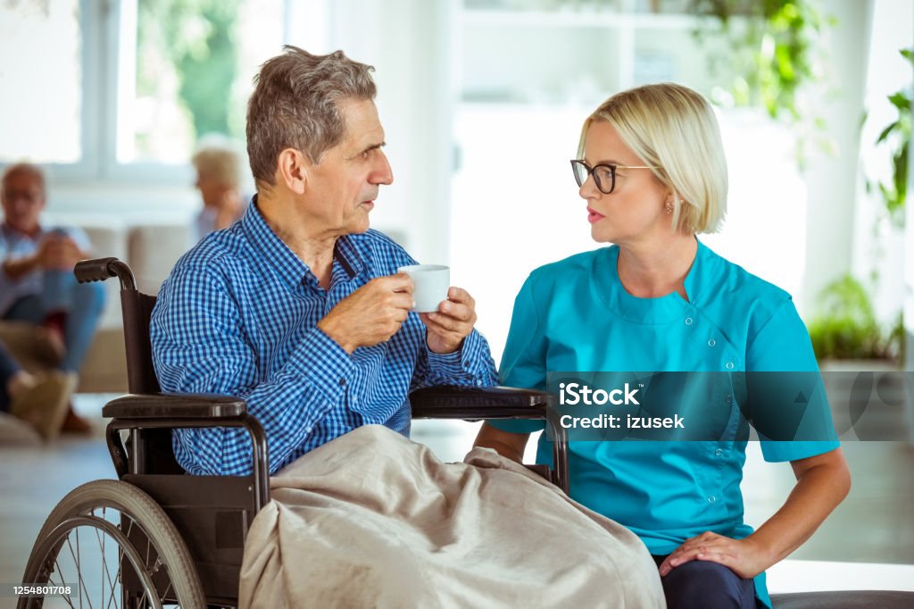 Nurse talking with senior man in retirement house Friendly home nurse talking senior man sitting in wheelchair. They are in nursing home. 70-79 Years Stock Photo