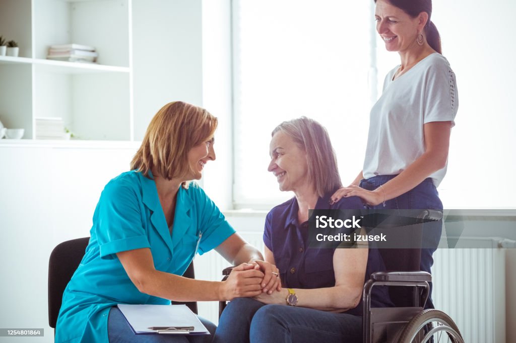 Nurse talking with smiling senior woman in hospital waiting room Nurse and elderly lady discussing medical test results. Senior woman sitting in wheelchair, social worker assisting her. Hospital Stock Photo