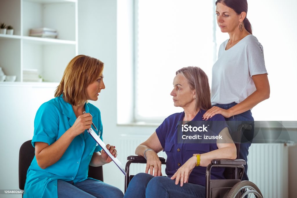 Nurse talking with worried senior woman in hospital waiting room Nurse and elderly lady discussing medical test results. Senior woman sitting in wheelchair, social worker assisting her. Nurse Stock Photo