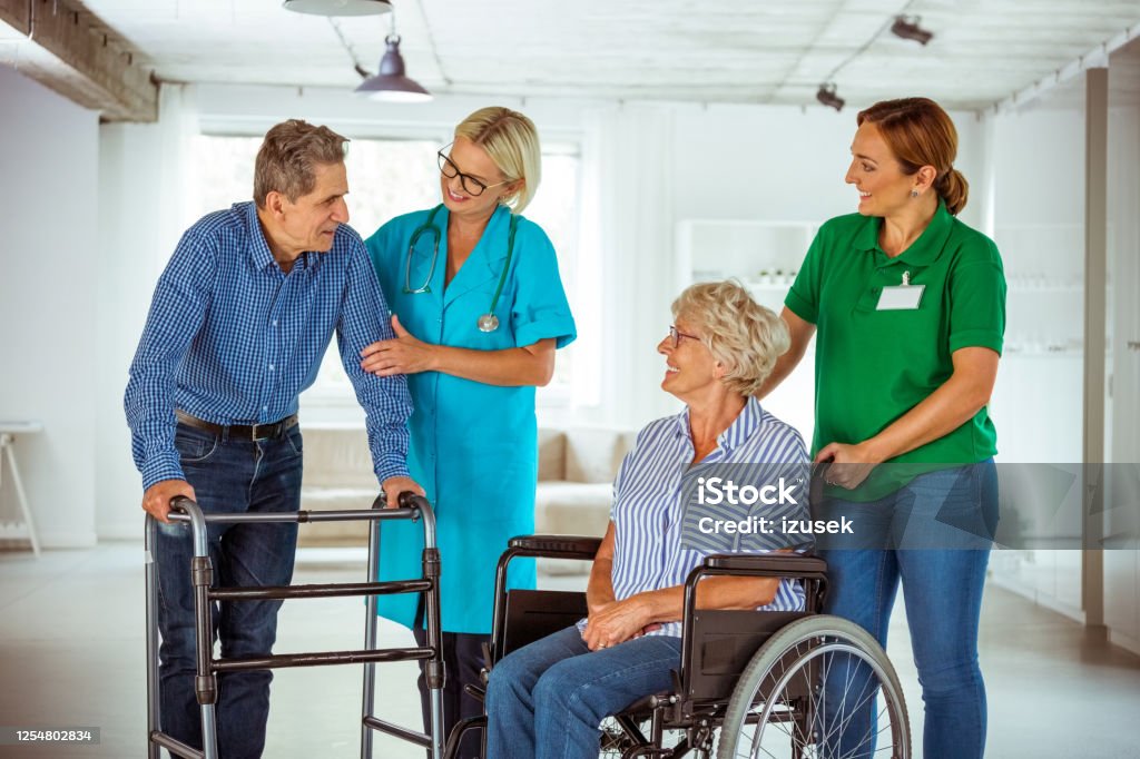 Senior people with nurses in retirement house Happy nurses and senior people in nursing home. Man using walking frame and elderly lady sitting in wheelchair. 70-79 Years Stock Photo