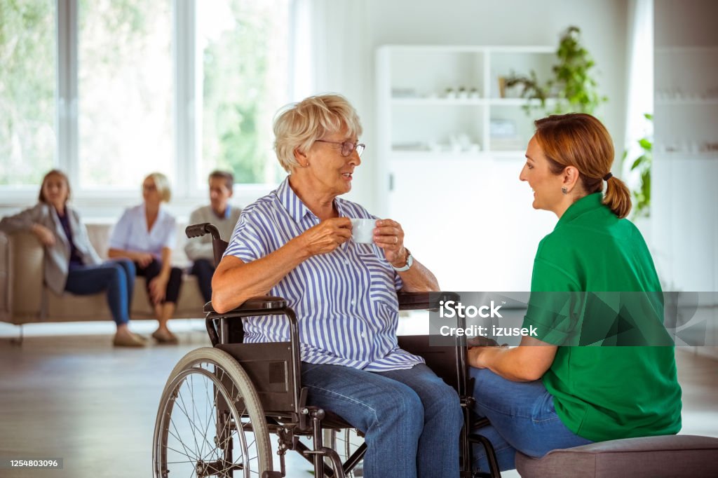 Senior woman in wheelchair talking with social worker Female social worker supporting senior woman. Elderly lady sitting in wheelchair in retirement house and drinking coffee. Happiness Stock Photo