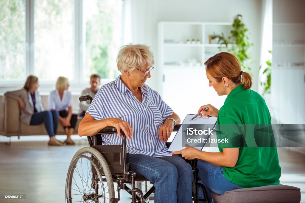 Senior woman in wheelchair talking with social worker Female social worker holding clipboard and explaining senior woman 
medical test results. Elderly lady sitting in wheelchair in retirement house. Rules Stock Photo