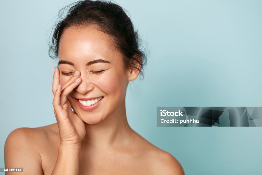 Beauty face. Smiling asian woman touching healthy skin portrait Beauty face. Smiling asian woman touching healthy skin portrait. Beautiful happy girl model with fresh glowing hydrated facial skin and natural makeup on blue background at studio. Skin care concept Women Stock Photo