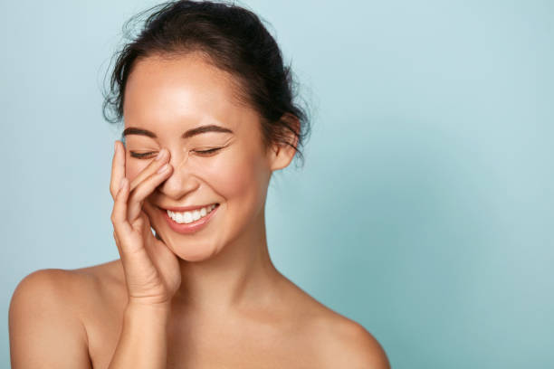 Beauty face. Smiling asian woman touching healthy skin portrait Beauty face. Smiling asian woman touching healthy skin portrait. Beautiful happy girl model with fresh glowing hydrated facial skin and natural makeup on blue background at studio. Skin care concept fresh faced stock pictures, royalty-free photos & images