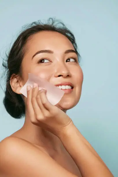 Face skin care. Smiling woman using facial oil blotting paper portrait. Closeup of beautiful happy asian girl model with natural makeup using oil absorbing sheets, beauty product at studio