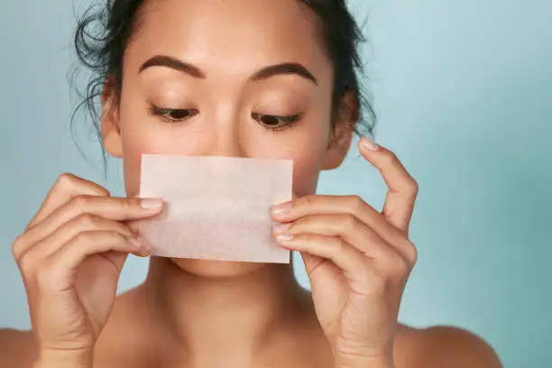 Skin care. Woman holding facial oil blotting paper portrait. Closeup of beautiful asian girl model with natural face makeup looking at oil absorbing tissue, beauty product.