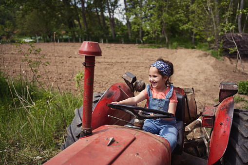 An adorable cute little blonde girl playing with a tractor toy sitting on rocks near a house