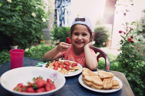 Cute little girl eating strawberries and waffles in her back yard during a warm spring morning