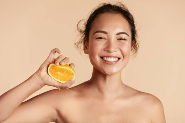 Beauty. Woman with radiant face skin squeezing orange portrait Beauty. Woman with radiant face skin squeezing orange in hand portrait. Beautiful smiling asian girl model with natural makeup, glowing facial skin and citrus fruit. Vitamin C cosmetics concept oranges with faces stock pictures, royalty-free photos & images