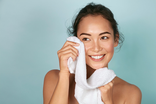 Washing face. Closeup of woman cleaning skin with towel portrait. Beautiful asian girl model wiping facial skin with soft facial towel, removing makeup. High quality studio shot