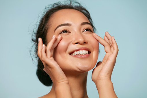 Skin care. Woman with beauty face touching healthy facial skin portrait. Beautiful smiling asian girl model with natural makeup touching glowing hydrated skin on blue background closeup