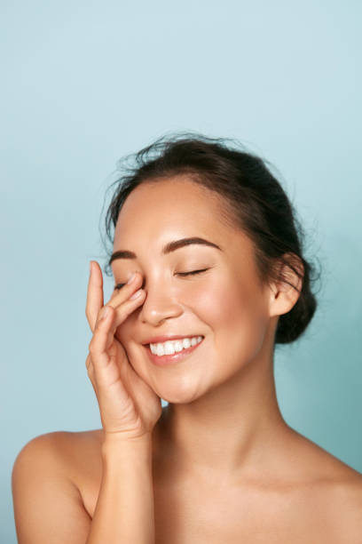 beauty face. smiling asian woman touching healthy skin portrait. beautiful happy girl model with fresh glowing hydrated facial skin and natural makeup on blue background at studio. skin care concept - women beauty innocence make up imagens e fotografias de stock