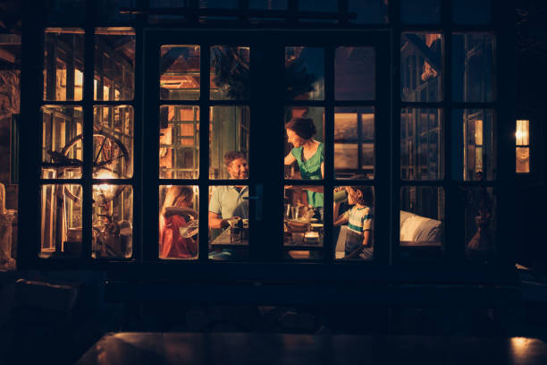 Happy family of four sitting at the dinning table in the dinning room, while woman sets dinner on the table. Photo is taken trough a window from outside of house.