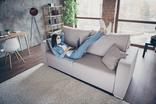 Nice attractive brunette girl lying on divan wearing safety mask using laptop, studying online courses watching lesson tutorial spending free time in modern loft industrial house flat apartment