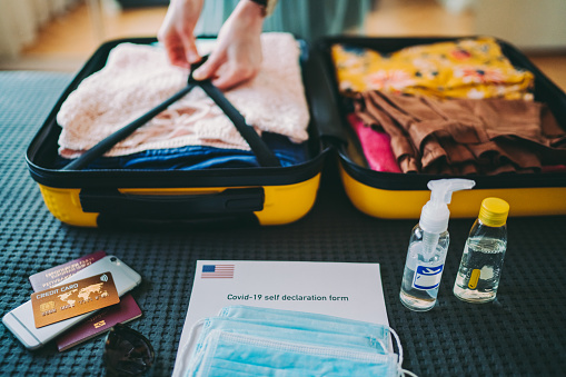Woman packing face masks and airplane travel-sized antibacterial hand gels for antiseptic hand washing during flight, also COVID-19 self-declaration form to be completed before landing
