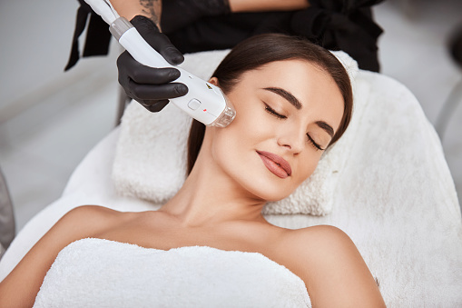beautiful girl lying in spa salon covering breast with white towel and getting massage for her cheek with professional apparat, lovely woman in beauty and spa concept