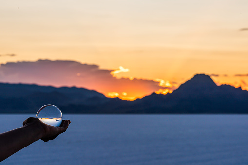 Person hand holding crystal ball view of round glass globe with reflection of Bonneville salt flats mountains at colorful sunset in Utah