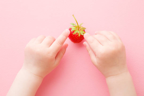 Baby fingers touching and exploring one red strawberry on light pink table background. Fresh berry. Pastel color. Closeup. Point of view shot. Top down view.