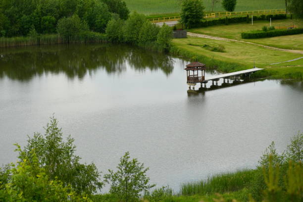 A view of a small lake or pond surrounded from all sides with dense forests, moors, and farmlands with a small wooden jetty or marina with a small platform leading to it located next to the coast vista de un pequeño lago o estanque rodeado por todos los lados con densos bosques, páramos y tierras de cultivo con un pequeño embarcadero o puerto deportivo de madera con una pequeña plataforma que conduce a él situado junto a la costa