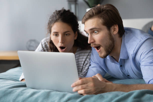 충격을 받은 젊은 부부는 노트북 화면을 보고, 침대에 누워 - mouth open retail shock surprise 뉴스 사진 이미지