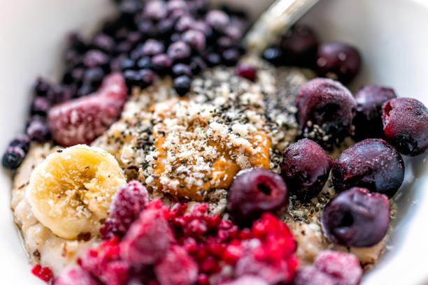 macro primo piano di ciotola di porridge d'avena con burro di arachidi, bacche congelate lamponi mirtilli e ciliegie con semi di banana e chia - oatmeal heat bowl breakfast foto e immagini stock