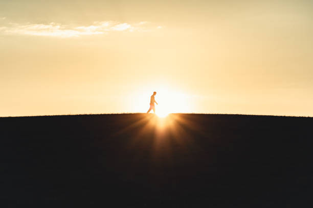Young man walking on top of the hill. Hike along footpath against the moody sunset sun. Backlit young man silhouette against the skyscape and hill towards the atmospheric horizon. Youth Outdoor Hiking Nature Moody Sunset Freedom Concept.
