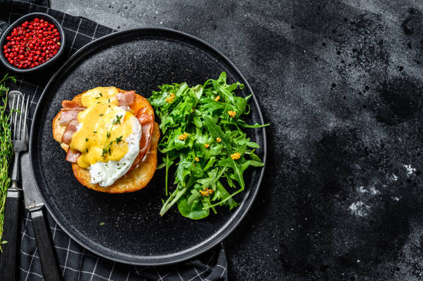 istock Brioche sandwich with bacon, egg Benedict and hollandaise sauce. Garnish with arugula salad. Black background. Top view. Copy space 1254894501