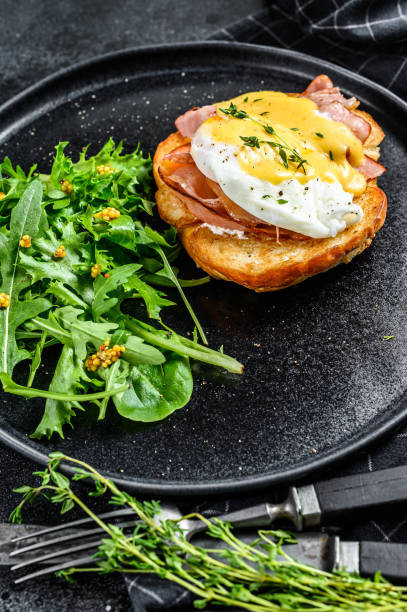 istock Brioche sandwich with bacon, egg Benedict and hollandaise sauce. Garnish with arugula salad. Black background. Top view 1254894505