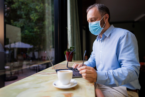 Mature Caucasian businessman with a protective face mask sitting at the coffee place and working on his laptop.