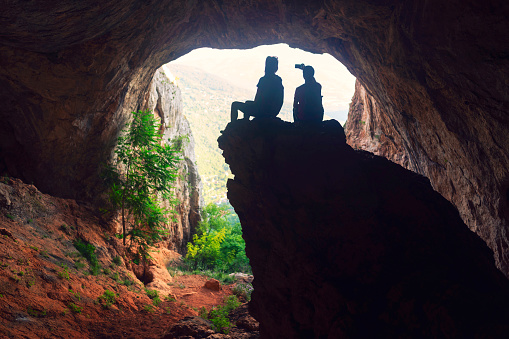 Young woman and young man in cave mountain sitting and looking at the view. Man using his phone to take a selfie.