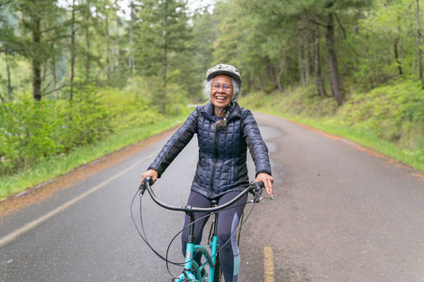 Senior woman riding bicycle on rural road An ethnic senior woman rides her bicycle along a country road in the forest. The road is sloping downhill as she rides toward the camera. active disruptagingcollection stock pictures, royalty-free photos & images