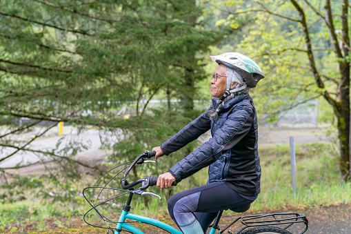 An ethnic senior woman happily rides a bicycle along a rural road on a summer afternoon. She is cycling from right to left down a slight incline and looking up with a content expression as she enjoys the beauty and solitude around her.