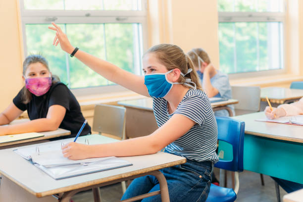 A thirteen year old girl is sitting at her desk in class with a mask to protect herself from Covid-19. A thirteen year old girl is sitting at her desk in class with others students around her. She is wearing a mask to protect herself from Covid-19. She is looking at the camera. The others students around her are working. teenage girls pretty smile looking at camera waist up stock pictures, royalty-free photos & images