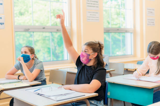 A thirteen year old girl is sitting at her desk in class with a mask to protect herself from Covid-19. A thirteen year old girl is sitting at her desk in class with others students around her. She is wearing a mask to protect herself from Covid-19. She is looking at the camera. The others students around her are working. teenage girls pretty smile looking at camera waist up stock pictures, royalty-free photos & images