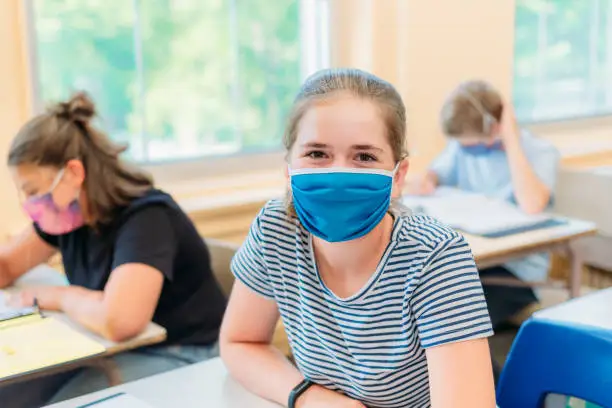 Photo of A thirteen year old girl is sitting at her desk in class with a mask to protect herself from Covid-19.