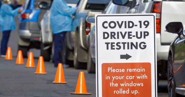 A "COVID-19 Drive-Up Testing" Sign Sits in the Foreground While Two Female Nurses Wearing Gowns and Surgical Face Masks Talk to Patients in their Cars in a Drive-Up (Drive Through) COVID-19 (Coronavirus) Testing Line Outside a Medical Clinic/Hospital Outd A "COVID-19 Drive-Up Testing" Sign Sits in the Foreground While Two Female Nurses Wearing Gowns and Surgical Face Masks Talk to Patients in their Cars in a Drive-Up (Drive Through) COVID-19 (Coronavirus) Testing Line Outside a Medical Clinic/Hospital Outdoors (Second Wave) in the Background drive through photos stock pictures, royalty-free photos & images