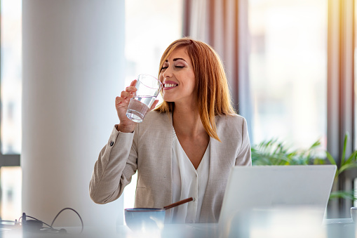 Businesswoman drinking water while working in her office. Close up of woman using a computer while holding a glass of water. Dehydrated female office worker drinking still mineral water