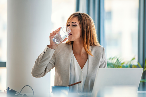 Office worker drinking water working on line. Young businesswoman drinking water in office. Happy beautiful young woman drinking water. Smiling female model holding transparent glass in her hand.