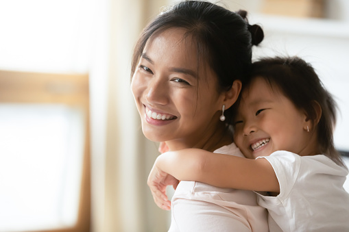 Adorable sweet little preschool kid daughter cuddling from back smiling beautiful vietnamese mother, enjoying sweet tender family moment together indoors, having fun at home, head shot close up.