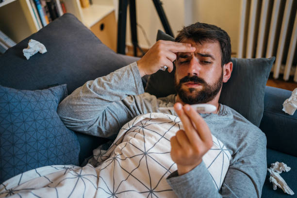 Sick man checking the temperature Sick man, wrapped in a blanket, is measuring a temperature while lying in bed. cold and flu man stock pictures, royalty-free photos & images