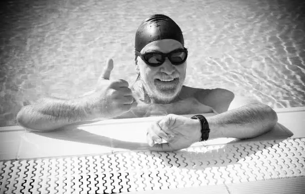 Happy senior man doing sport in the outdoor swimming pool checking the time of the swim. Black swim cap and goggles. Concept of active elderly people during retirement